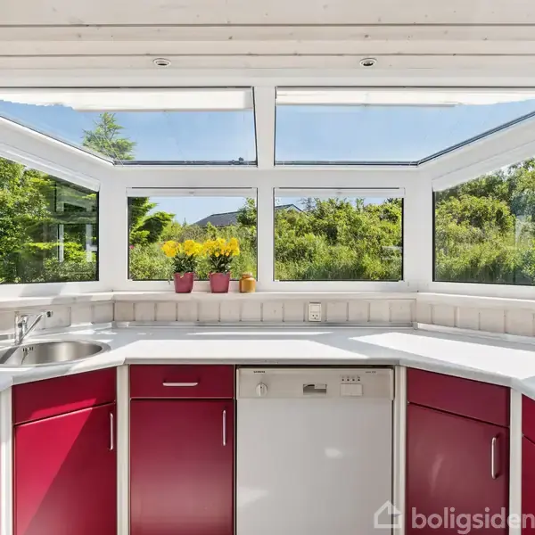 Et køkken med pink skabe omringer en hvid køkkenbordplade med vask og opvaskemaskine. Vinduer med udsigt til grøn have. Tre gule blomster i potter står på vindueskarmen.