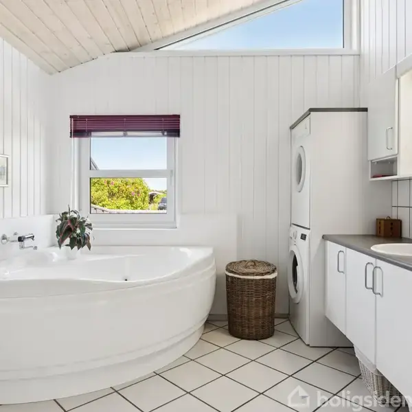 Et stort, lyst badeværelse med et hjørnebadekar, en vaskemaskine og tørretumbler stabel, en vask med skab og spejl og en vasketøjskurv. Vindue med udsigt til træer udenfor.