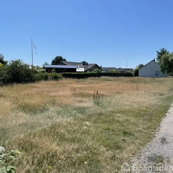 En åben, græsbeklædt grund ligger stille under en klar blå himmel. Til højre er en grussti, og i baggrunden ses et afgrænset boligområde med flere huse og træer.