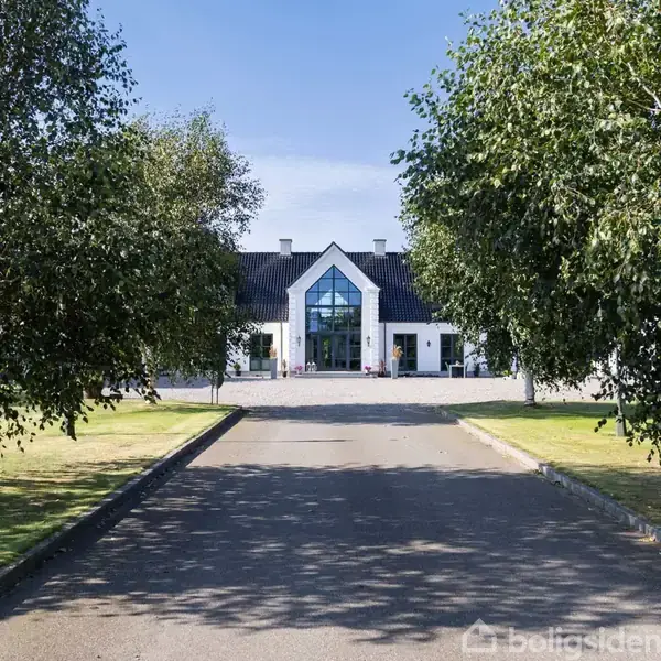 En stor, moderne hvid villa med glasfacade står for enden af en træomkranset indkørsel. Omkringliggende grønt græsplæne skaber en indbydende atmosfære.