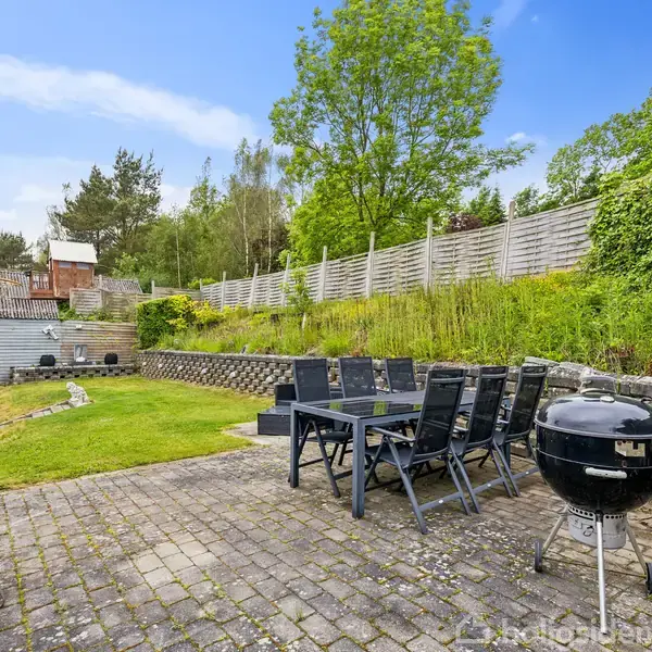 Grill og spisebord står på en terrasse af sten i en have med høje træer og græs, omgivet af en stakit og en stenmuret hævet bed.