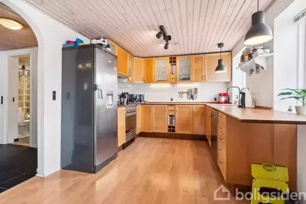 Køkken med træskabe rummet langs væggene, grå køleskab til venstre. Rummet har lyse trægulve, hvide vægge og loft af træ. Der er pendellamper og køkkenudstyr på bordpladen.