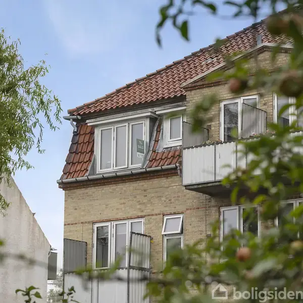 En murstensbygning med rødt tegltag, som har åbne vinduer og en lille altan. Omgivet af træer med modne æbler, under en klar blå himmel.