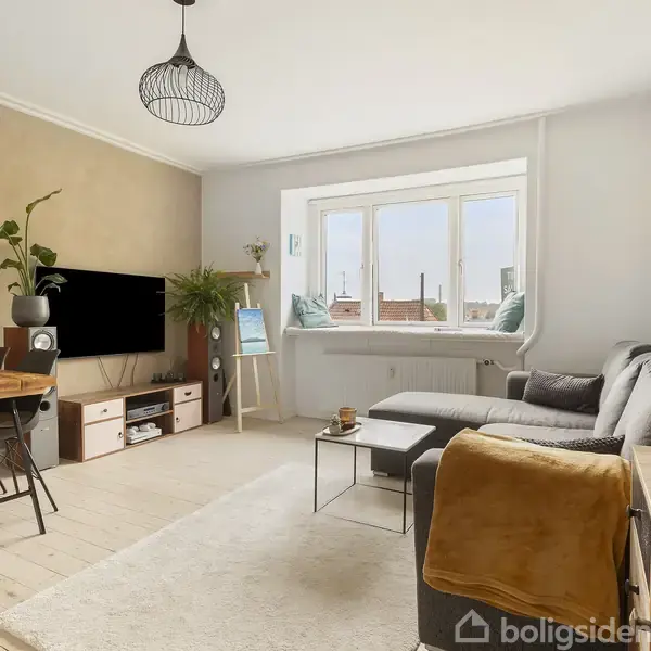 Stuen har en grå L-formet sofa foran et vindue med udsigt til rødtage. Et træbord står foran sofaen, mens en tv-reol og spisebord med stole er på den ene væg.