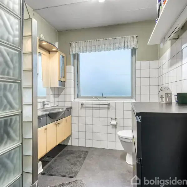 Badeværelse med toilet og vask, placeret mod en væg af hvide fliser. Et vindue er centralt på væggen, omgivet af en hylde, spejl, og opbevaringsskabe. Ingen tekst synlig.