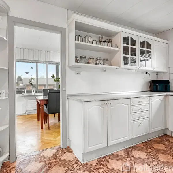 Et lyst køkken med hvide skabe og hylder indeholder service. En døråbning fører til en stue med store vinduer. Mønstret gulv og et lille vindue fuldender rummet.