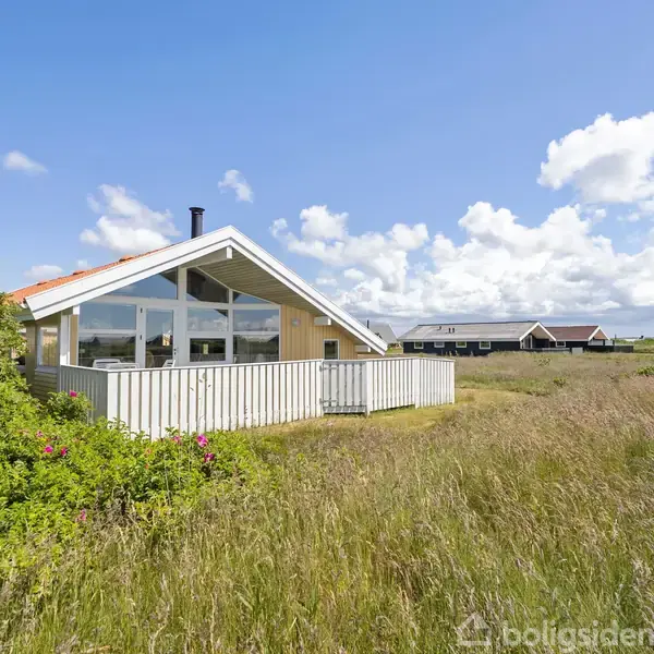 Sommerhus med store vinduer står stille. Omgivet af høj græs og blå himmel med enkelte skyer. Øvrige bygninger ses i baggrunden.