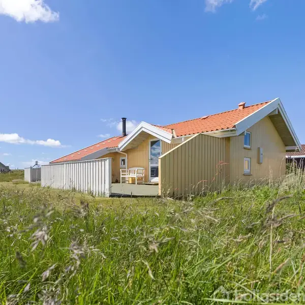 Gult sommerhus med rødt tag står på en græsmark. Der er blå himmel med få hvide skyer i baggrunden. Terrasse med stole foran huset.