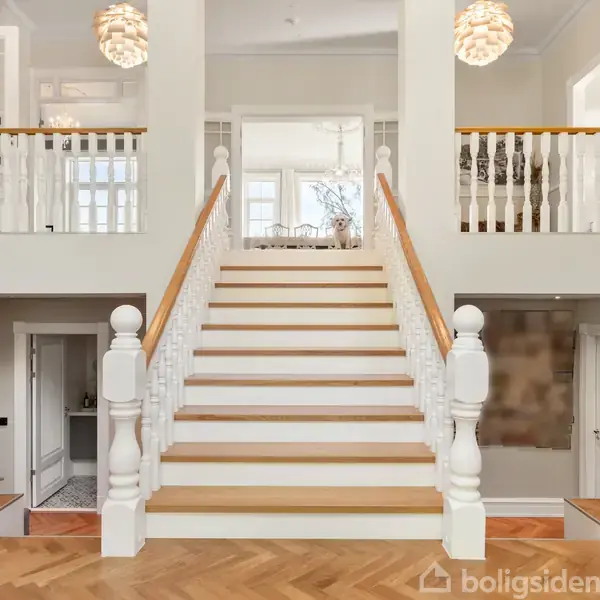 Trappe opad med hvide balustre og trægelænder, fører til rummelig øverste etage. Tofløjede døre og elegante lamper ses i elegant entré med parketgulv.
