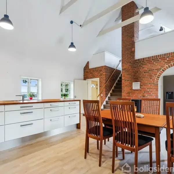 Et spisebord med seks stole står i et moderne køkken med hvide skabe. I baggrunden er der en murstensvæg med en døråbning, en trappe og ophængte lamper.