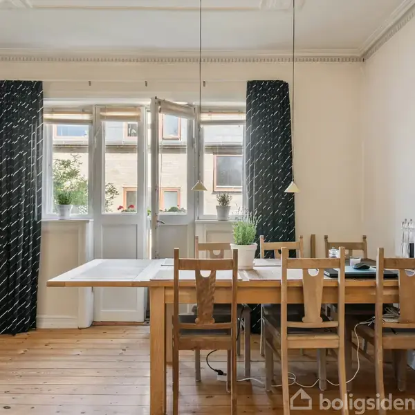 Et spisebord med seks træstole står i et lyst rum. Vinduer med mørke gardiner i baggrunden. En bordplante og nogle små genstande er placeret på bordet.