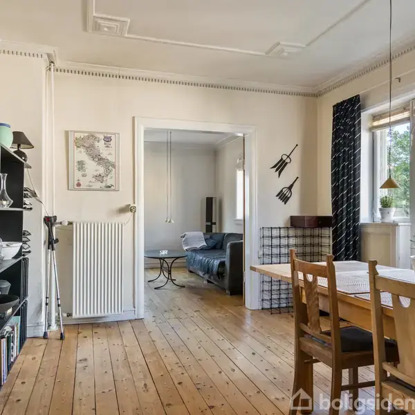 Spisestue med træbord og stole står ved store vinduer med udsigt til have. Til venstre, sort reolsystem med diverse pyntegenstande. I baggrunden ses en stue med sofa.