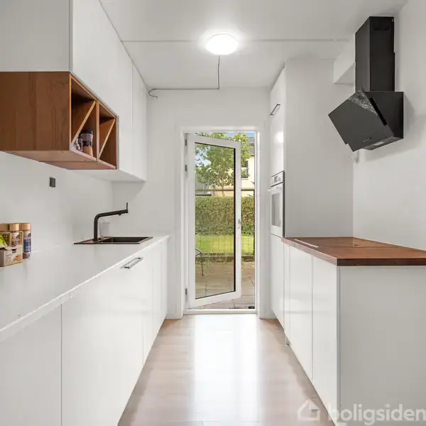 Et moderne køkken med hvide skabe og træbordplade. Kanden og opvaskemiddel er til venstre, mens en udgang til en have med grønne planter er i baggrunden.