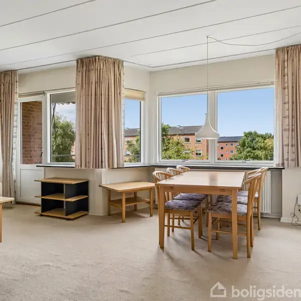 Stue med spisebord og stole. Ved siden af står en sofa og et lille bord. Store vinduer med gardiner slipper lys ind. Udsigt til andre bygninger udenfor.