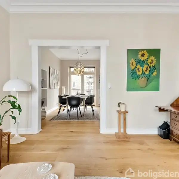 Stue med træmøbler og en plante på gulvet. I baggrunden ses et spiseområde med sort bord og stole. På væggen hænger et maleri af solsikker.
