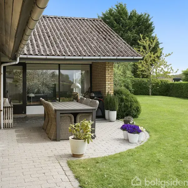 Terrasse med flettede havemøbler står på flisebelagt område, omgivet af velplejet græsplæne og buske. En murstensvæg med store vinduer afslører husets facade.