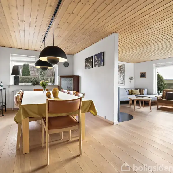 Spisebord i lyst træ med stole og gul dug, står under sorte lamper. Ved siden af er en opholdsstue med sofaer, vinduer og træloft skaber en lys atmosfære.
