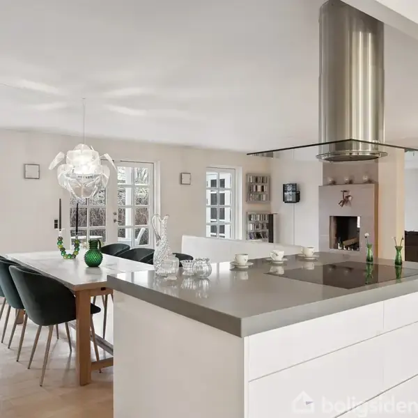 Køkkenø med glat bordplade, omgivet af barstole, står i et lyst køkken-alrum. Spisebord og stole er placeret ved siden af, mens en skorstensindsats ses i baggrunden med sofa.