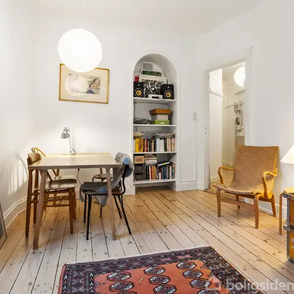 Et spisebord med stole står i et lyst værelse. En hvid hængelampe lyser rummet op. Bogreol og lænestol tilføjer atmosfære; en rød orientalsk tæppe pryder gulvet.