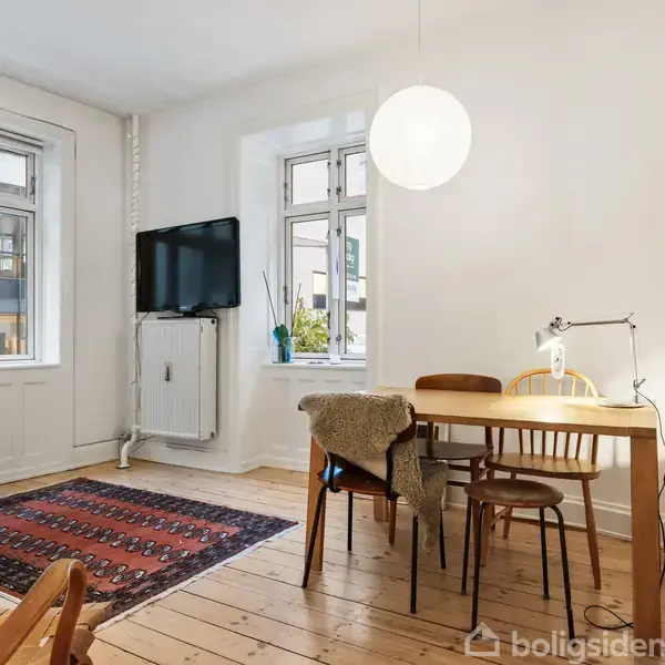 Et spisebord med stole står i et lyst værelse. En lampe hænger over bordet, og et tæppe ligger på gulvet. En fladskærm er monteret på væggen.