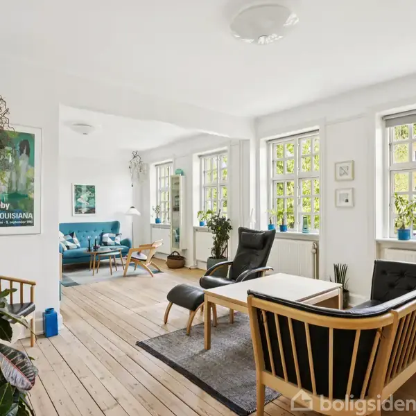 Stue med lyse vægge og trægulv indeholder sorte lænestole og sofa. Store vinduer lader lys strømme ind. Plakat på væggen viser "Per Kirkeby Louisiana". Planter og reoler skaber hyggelig atmosfære.