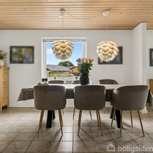Et spisebord med grå stole står i et lyst rum med flisegulv. Over bordet hænger to dekorative lamper. I baggrunden ses et vindue med udsigt til et hus og træer.