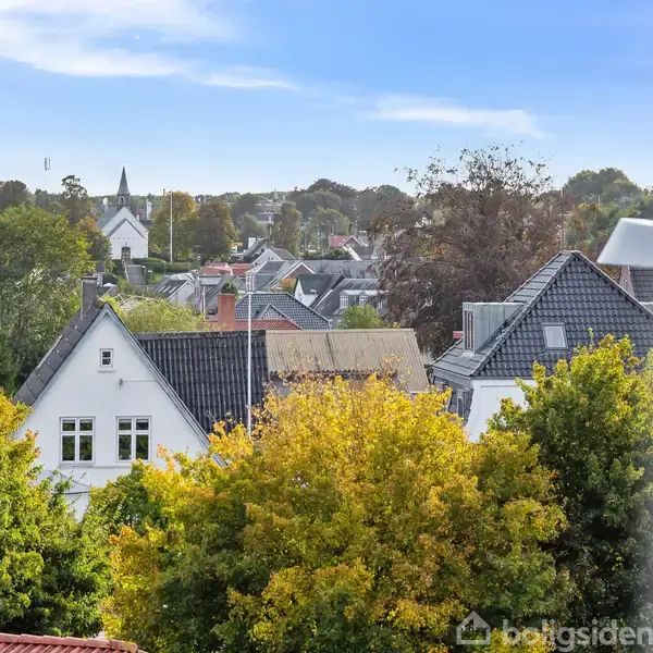 Flere huse med skrå tage og vinduer ses blandt træer med efterårsfarver. I baggrunden er en kirketårn og en blå himmel synlig, der skaber en fredfyldt atmosfære.