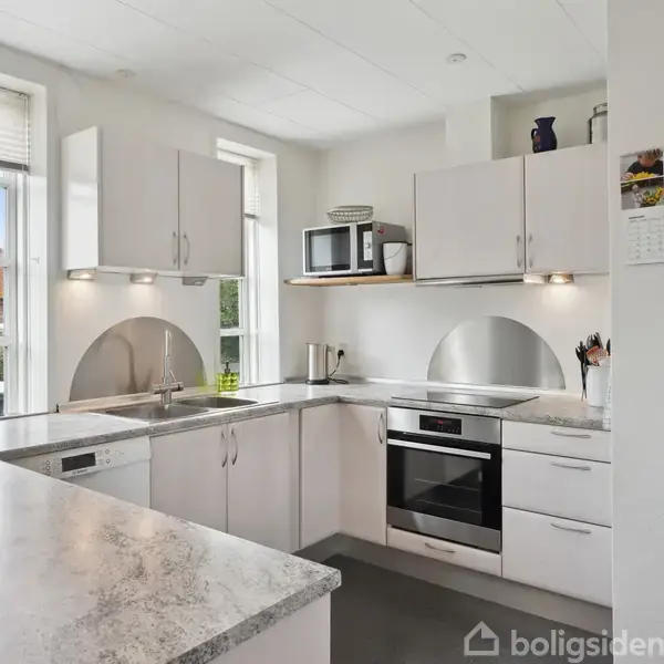 Et hvidt, moderne køkken med granitbordplade. Ovn og mikroovn er placeret indbygget i skabet. Der er vinduer med udsigt til huse, og en kalender hænger på væggen.