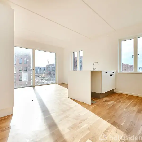 Et lyst rum med trægulv indeholder et moderne køkken med en hvid køkkenø. Store vinduer til venstre giver udsigt til en bygningsplads udenfor.