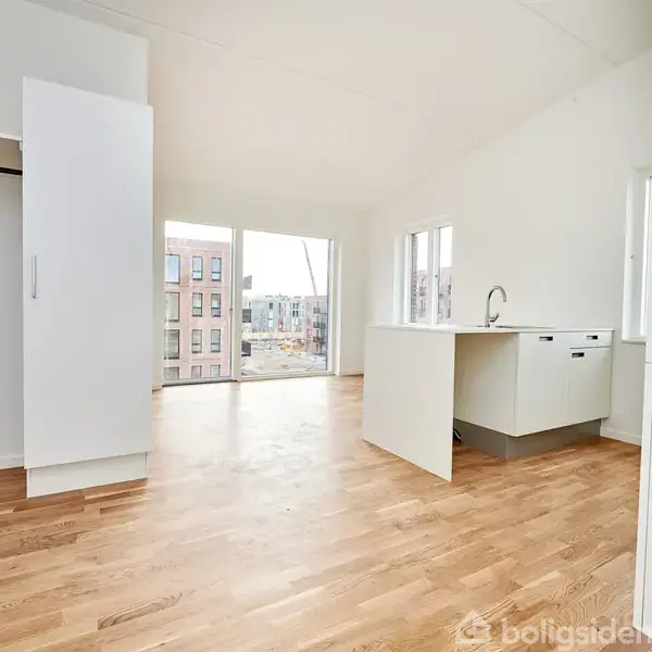 Et lyst, tomt rum med trægulv har et lille køkken med en vask. Store vinduer i baggrunden giver udsigt til bygninger udenfor. Hvidt skab langs væggen.