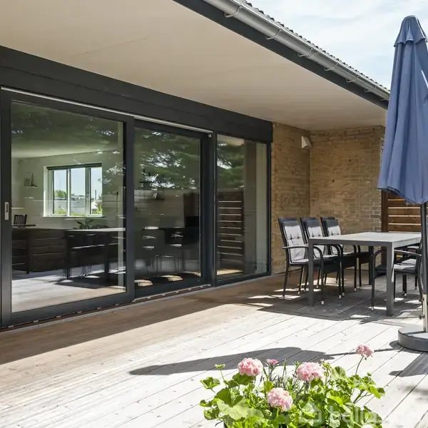 Terrassebord med seks stole står på en rummelig træterrasse, hvor en blå parasol er slået op. Store glasskydedøre fører indtil huset, og blomster pryder forgrunden.