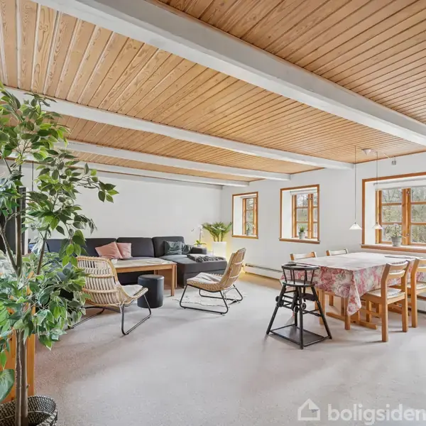 Stue indrettet med sort sofa og stole omkring et sofabord, placeret på et lyst gulv. Ved siden af er et spisebord med stole. Vinduerne slipper naturligt lys ind i rummet.