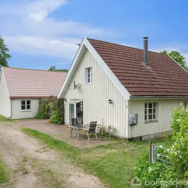 Et hvidt træhus med rødt tag har en terrasse foran med to stole og et bord. En grusvej løber langs huset, omgivet af grønt løv og en klar blå himmel.
