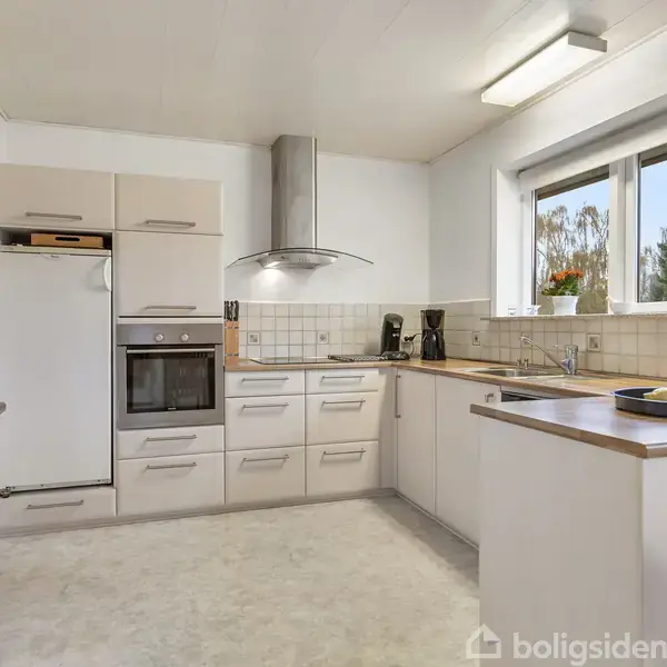 Køkken med hvide skabe og stålhåndtag, står ryddeligt. Komfur og ovn indbygget mellem skabe. Stort vindue med udsigt til træer udenfor giver naturligt lys.