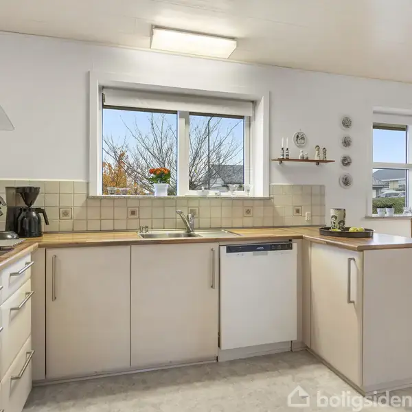 Et lyst køkken med hvide skabe, en indbygget ovn og en opvaskemaskine. Kaffeapparat og planter står på bordpladen. I baggrunden er der et spisebord ved vinduet.