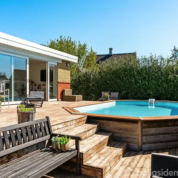 Swimmingpool i træterrasse, placeret i en solrig have. Omkranset af træer og buske. Terrasse med stole og bord udenfor et moderne hus med store vinduer.