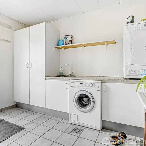 Vaskemaskine placeres under arbejdsbord i vaskerum med hvide skabe. Ovenpå bordet står en tørretumbler. Rummet har flisegulv, en måtte, grønne planter og en glasdør mod haven.