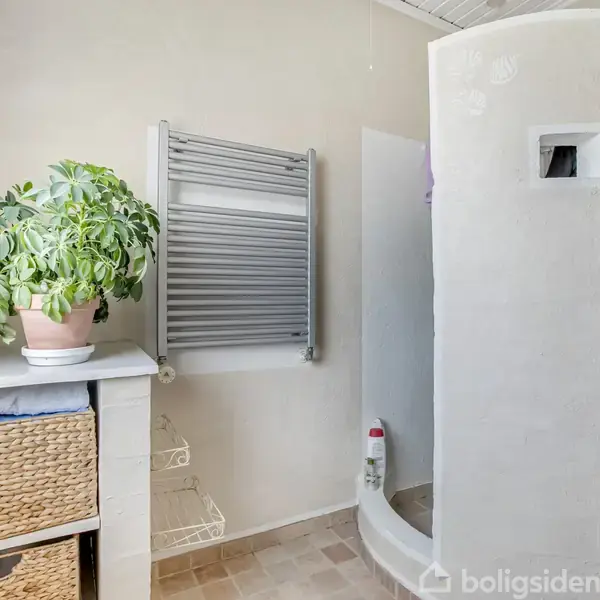 En cirkulær bruseniche står i et lyst badeværelse med beige fliser. En grøn plante i en potte står ovenpå en hylde med flettede kurve, mens et håndklædetørrer hænger på væggen.