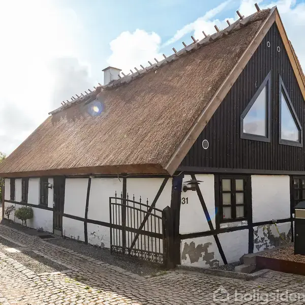 Stråtækt hus med bindingsværk står stille ved en brostenssti, omgivet af træer og buske. Nummer 31 på facaden ved siden af en sort smedejernslåge.