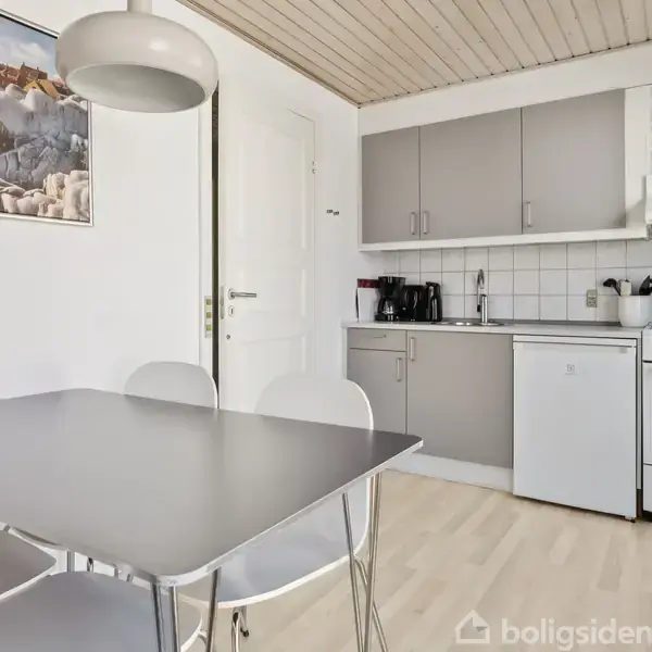 Bord med stole står i centrum, omgivet af et moderne køkken i lyse farver. Der hænger en lampe over bordet og et naturmaleri på væggen ved siden af døren.