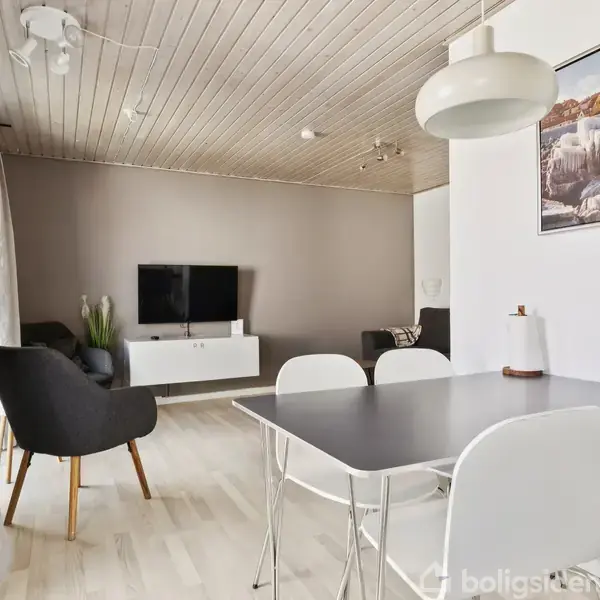 Et moderne stueområde med sort bord og hvide stole. Et TV er monteret på væggen over en hvid skænk. Rummet har lyst trægulv og en ophængt plakat på væggen.
