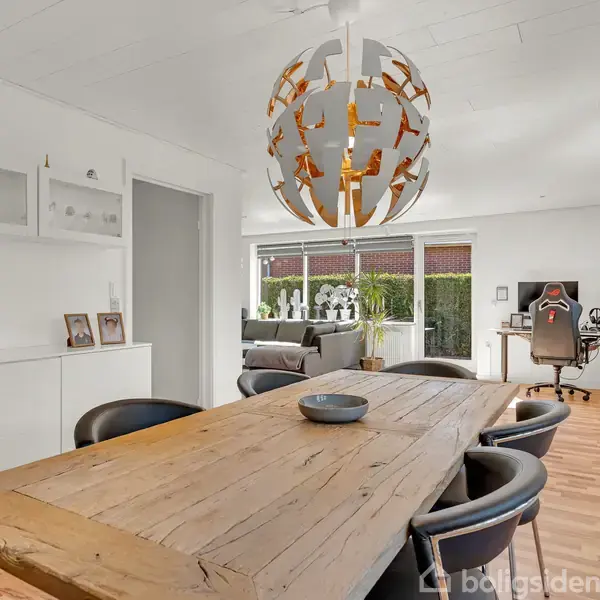 Rustikt spisebord står i midten af en stilren stue med hvide vægge, hvor lampen over bordet hænger. I baggrunden ses en kontorplads ved store vinduer, der giver udsigt til haven.