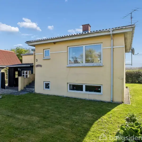 Et gult hus med store vinduer står i en have med grøn græsplæne. Til venstre er en carport med rødt tag. Huset har husnummeret 3.