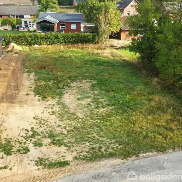 Tomt med ujævnt græsdække strækker sig mellem en murstensbygning til venstre og tæt beplantede træer til højre, i et hyggeligt byområde med flere huse i baggrunden.