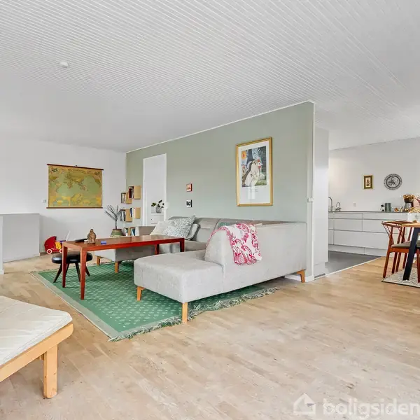 En lys stue med sofa og sofabord på et grønt tæppe, hvor en væg deler rummet fra køkken/spisestue-området med hvide møbler og spisebord i baggrunden.