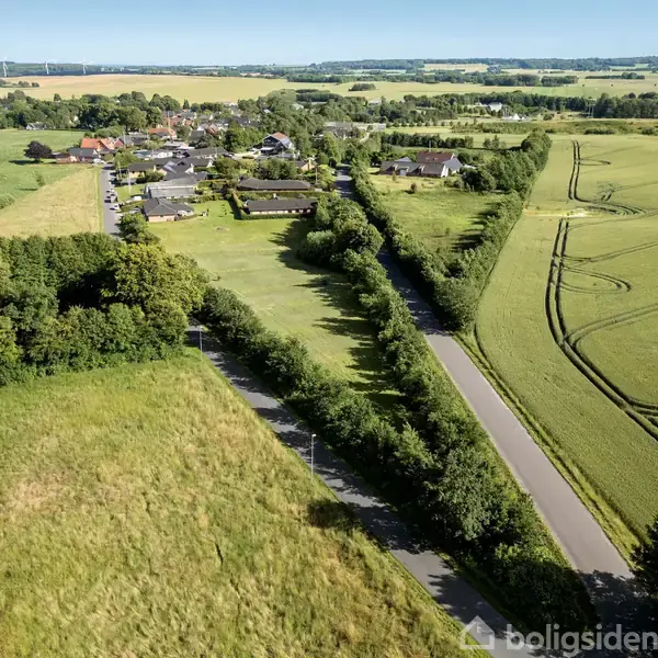 Luftfoto af en landsby med huse omkranset af skove og marker. En lang vej løber gennem landskabet, omgivet af grønne marker og spredte træer under en klar blå himmel.