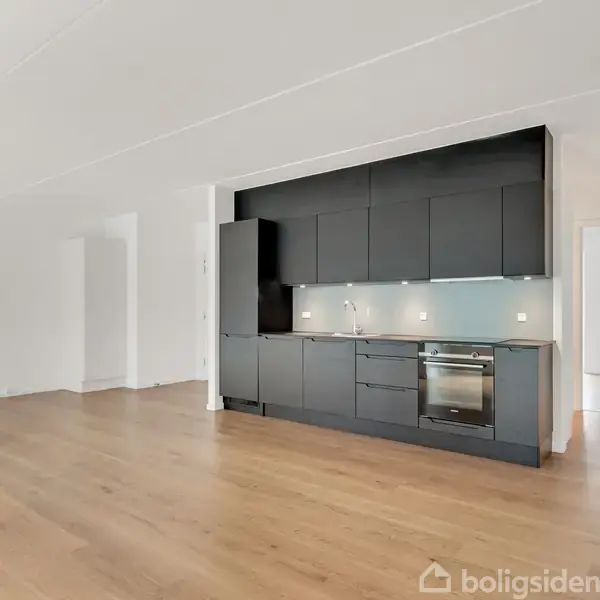 Køkken i et lyst rum; sorte skabe står langs væggen med indbygget ovn og vask; trægulv og hvidt loft. Der er en åben dør i siden, som fører til et andet rum.