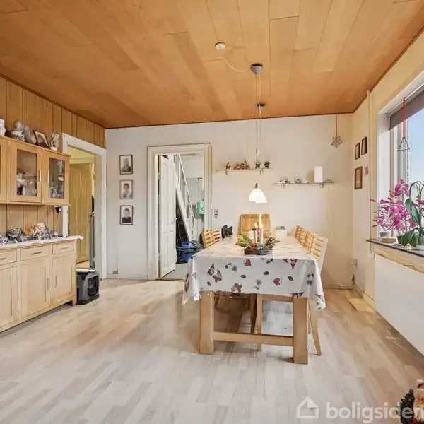 Et spisebord med dug er omgivet af seks træstole. Bordet har festlig dekoration. Til højre er et vindue med orkideer. Til venstre står et skab med udstillet porcelæn.