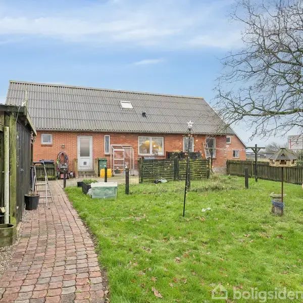 Rødt murstenshus set fra haven, med flisebelagt sti mod en græsplæne. Baggrunden viser et stort træ og diverse haveobjekter. Stakittet omkranser området.