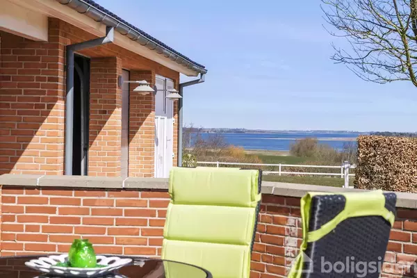 En terrasse med grønne stole står foran et hus med murstensvæg i solskin. I baggrunden ses et smukt landskab og havudsigt.